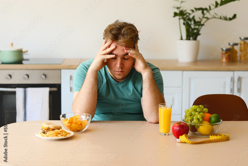 Upset young overweight man sitting at table with food in kitchen