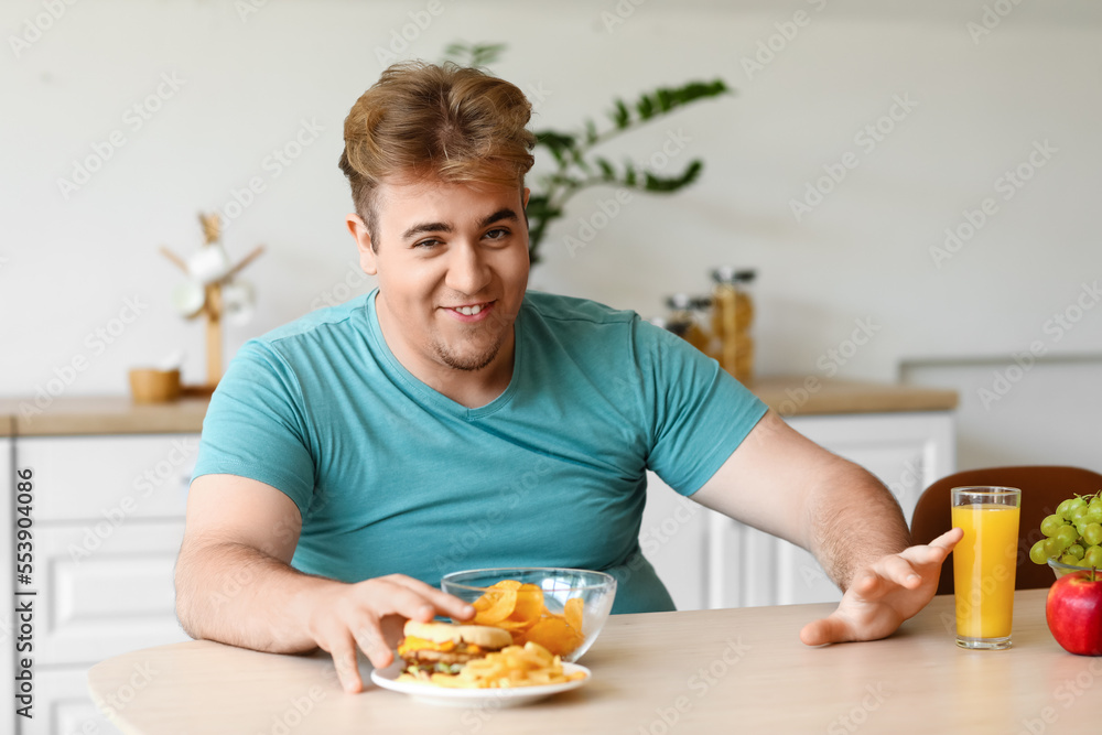 Young overweight man rejecting healthy food at table in kitchen