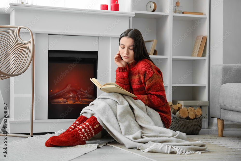 Young woman in warm sweater reading book near fireplace at home