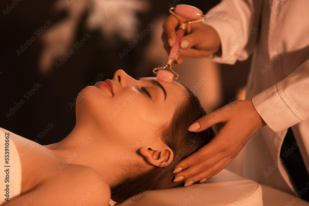 Pretty young woman receiving face massage with roller in beauty salon