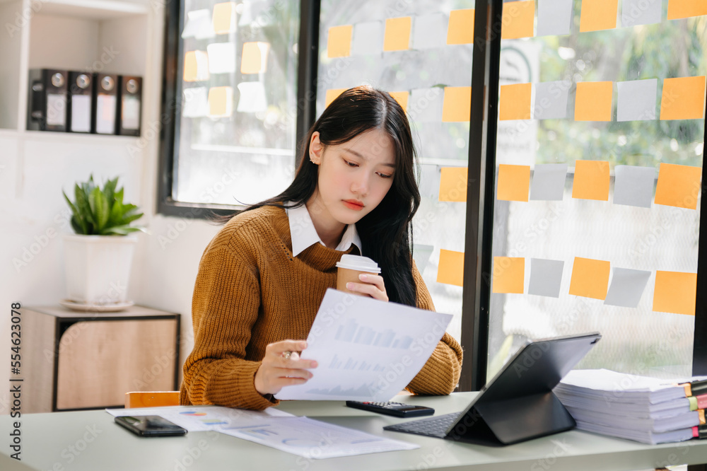 Confident beautiful Asian businesswoman typing laptop computer and digital tablet while holding coff