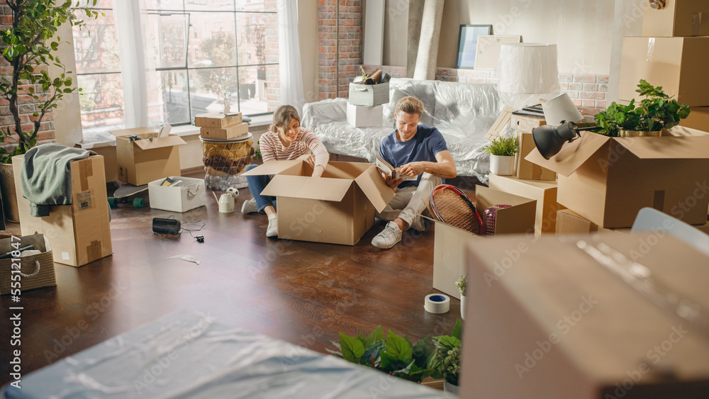 Happy Homeowners Moving In: Lovely Couple Sitting on the Floor of Cozy Apartment Unpacking Cardboard