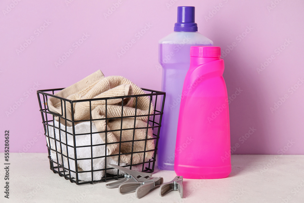 Laundry detergents and basket with clothes on table against lilac background