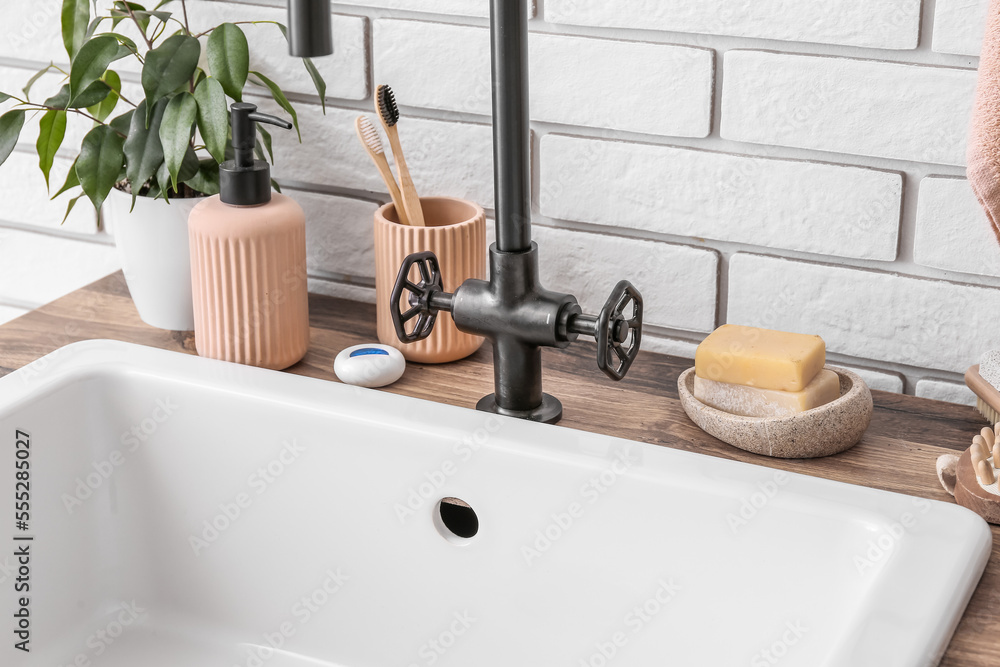 Table with ceramic sink, bath accessories and houseplant near white brick wall, closeup