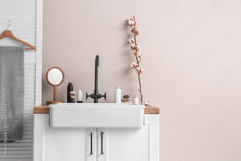 Table with ceramic sink, bath accessories and cotton flowers near light wall