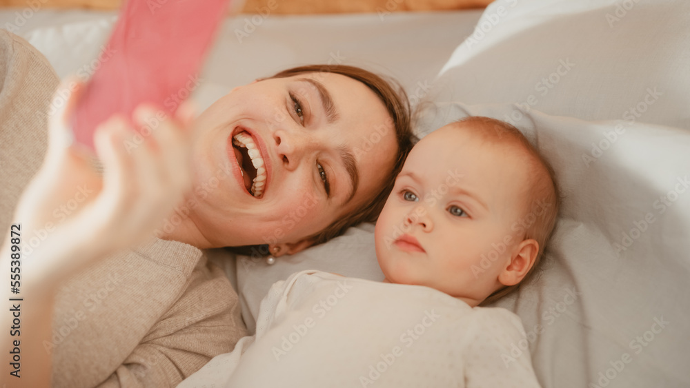 Young Beautiful Mother Lying in a Bed with Her Baby Girl, Making a Video Call on Smartphone. Having 