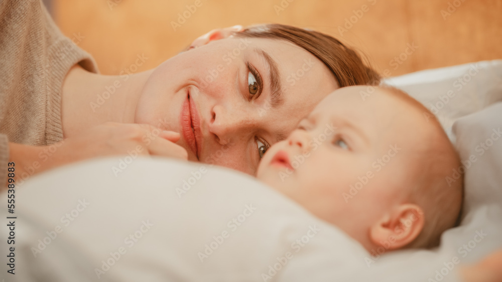 Loving Young Beautiful Mother Playing and Soothing Adorable Baby in Bed. Smiling Caring Mom and Cute