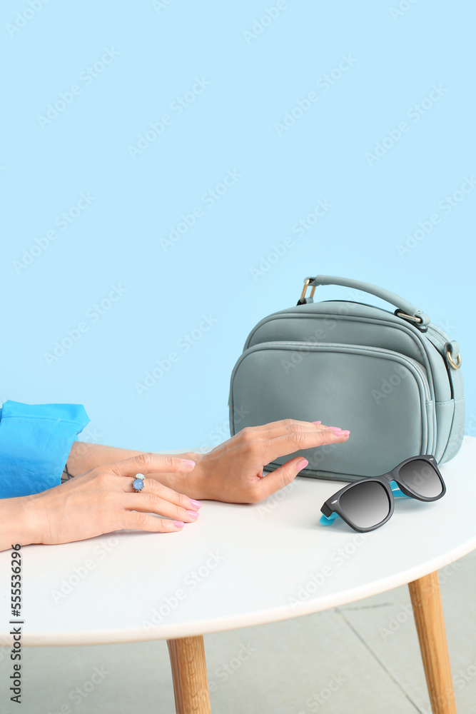 Female hands with stylish jewelry, handbag and sunglasses on white table against color background