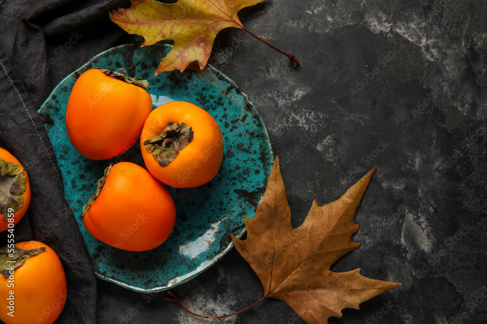 Plate with ripe persimmons and autumn leaves on dark background