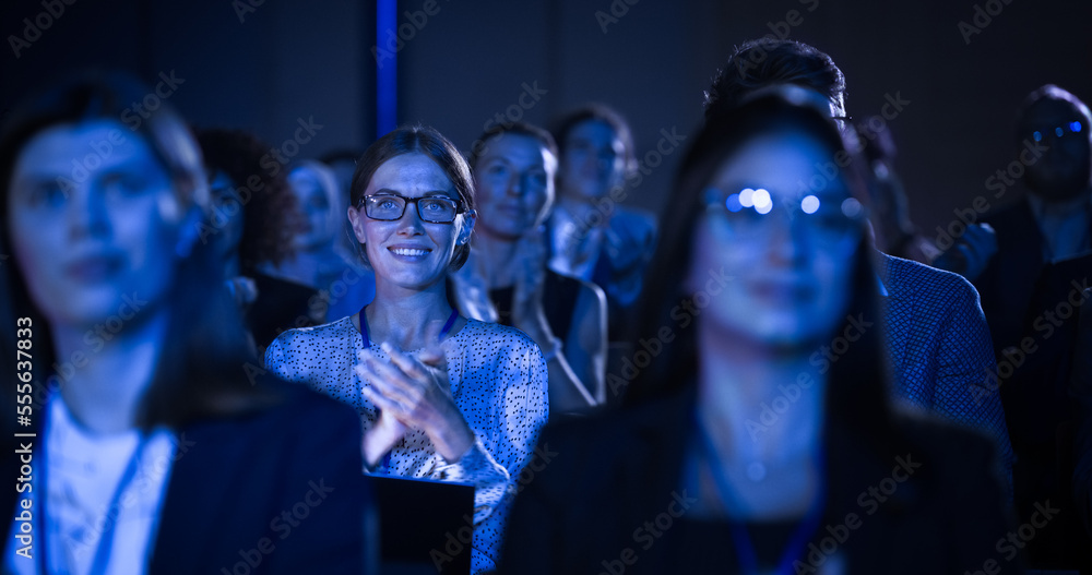 一位美丽的女性坐在会议上黑暗拥挤的礼堂里。一位年轻女性在鼓掌