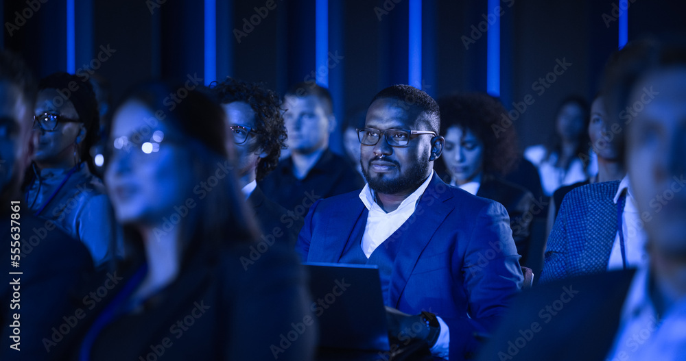 African Male Sitting in a Dark Crowded Auditorium at a Tech Conference. Professional Using a Laptop 