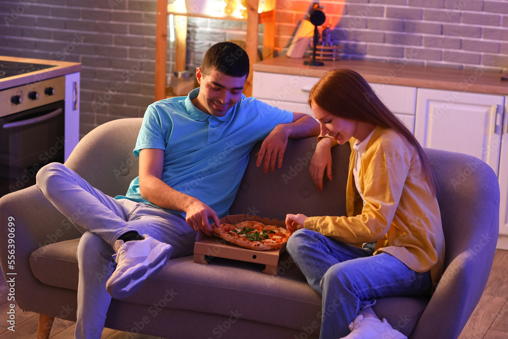 Young couple eating tasty pizza on sofa in kitchen at night