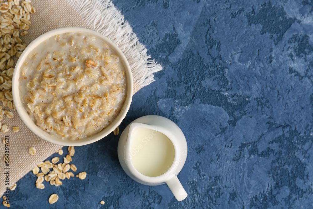 Bowl of tasty oatmeal, pitcher with milk, scattered flakes and napkin on blue table