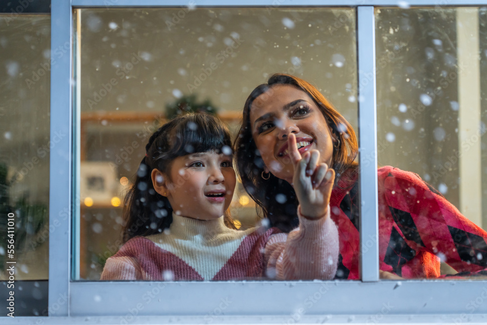 Adorable child looking at the window and admire snowflakes with mother. 