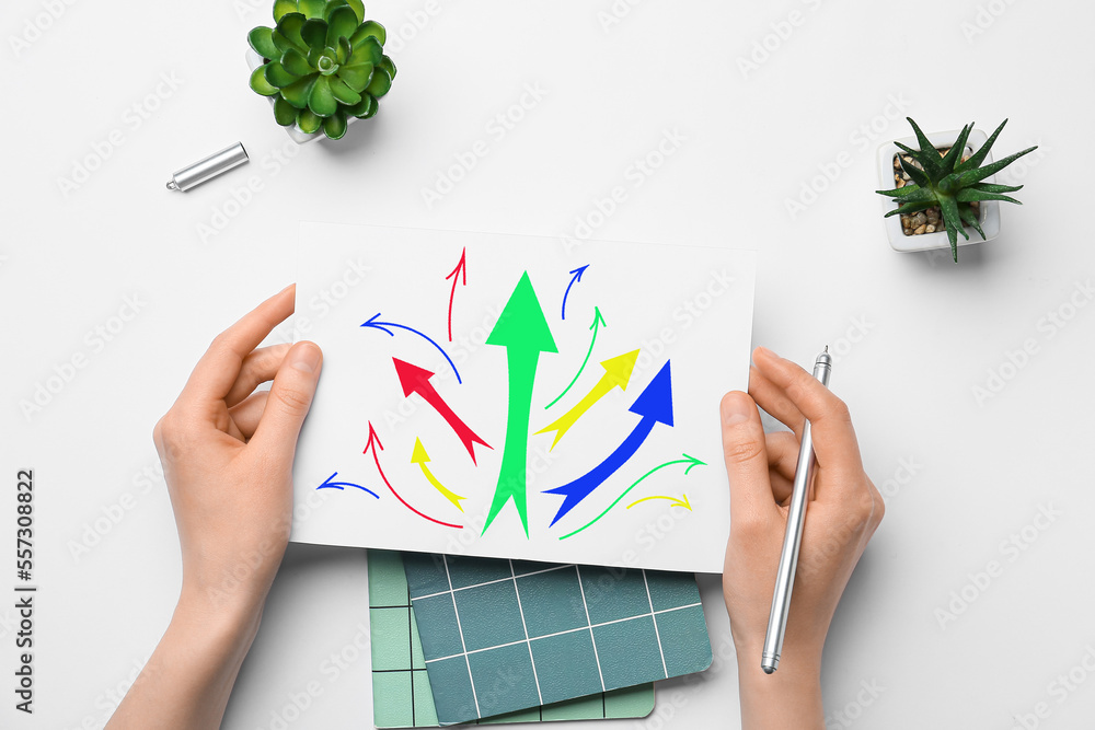 Woman holding paper sheet with drawn arrows and pen at office table, top view. Concept of choice