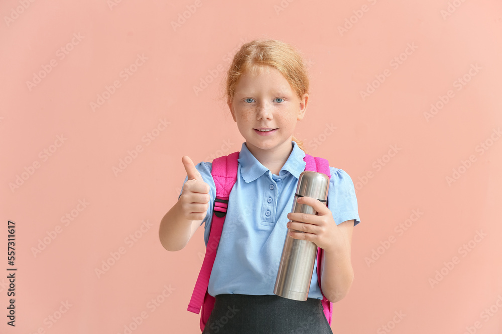 Little redhead schoolgirl with thermos showing thumb-up on color background