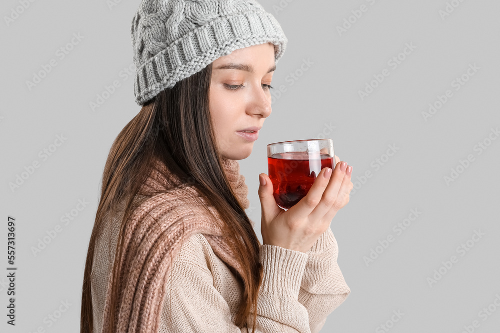 Young woman in warm hat with glass cup of tea on grey background, closeup