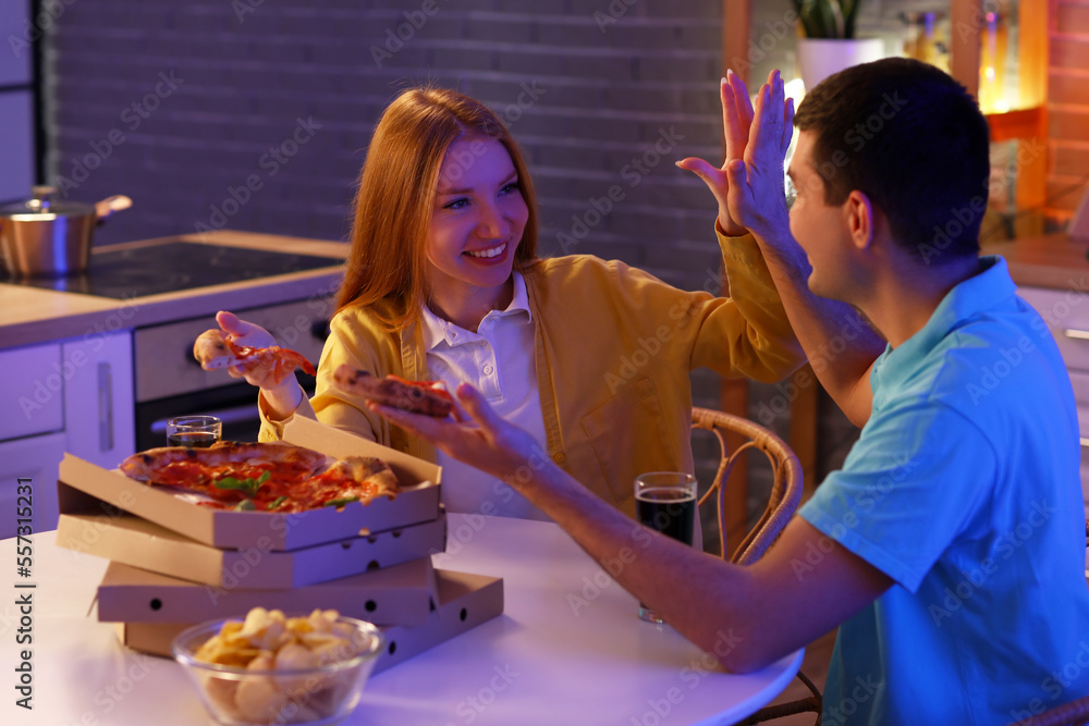 Young couple with pizza giving each other high-five in kitchen at night