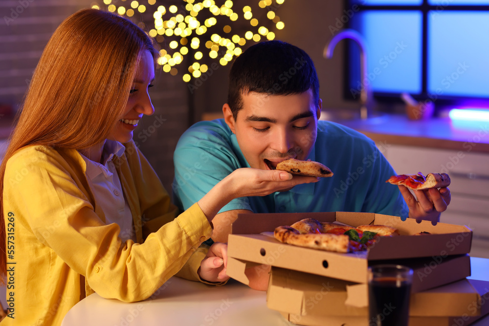 Young woman feeding her boyfriend with tasty pizza in kitchen at night