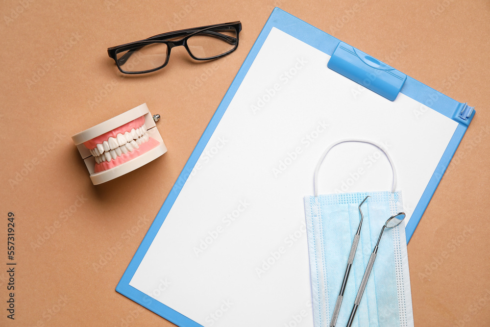 Clipboard with medical mask, dental tools, jaw model and eyeglasses on brown background