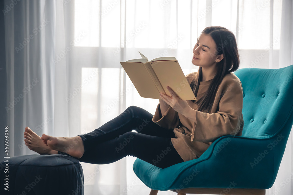 A young woman is reading a book while sitting on an armchair by the window.