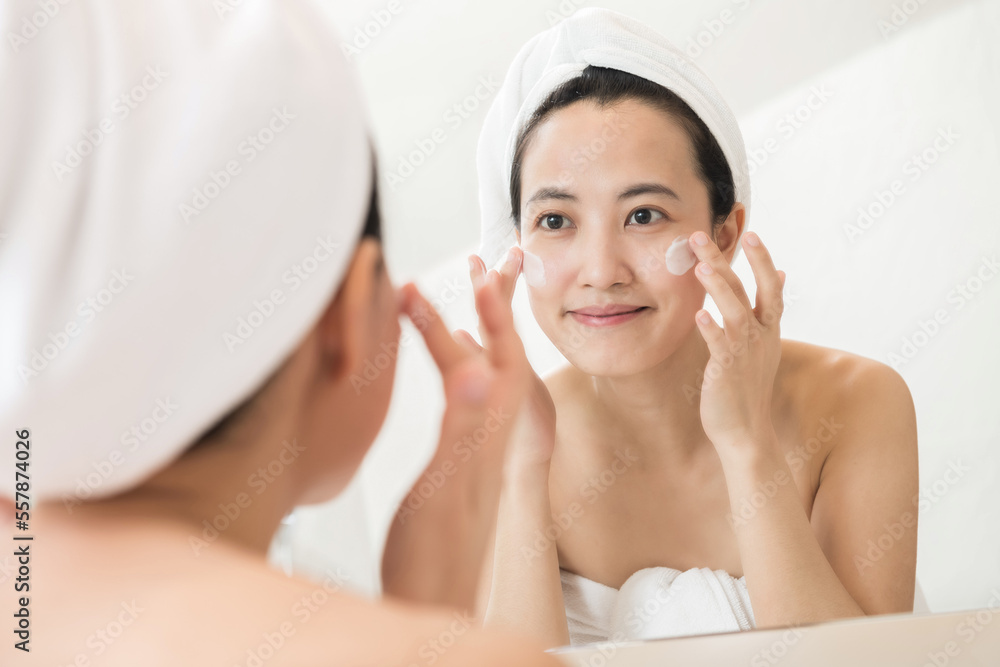 Happy young Asian woman applying face lotions while wearing a towel and touching her face in bathroo