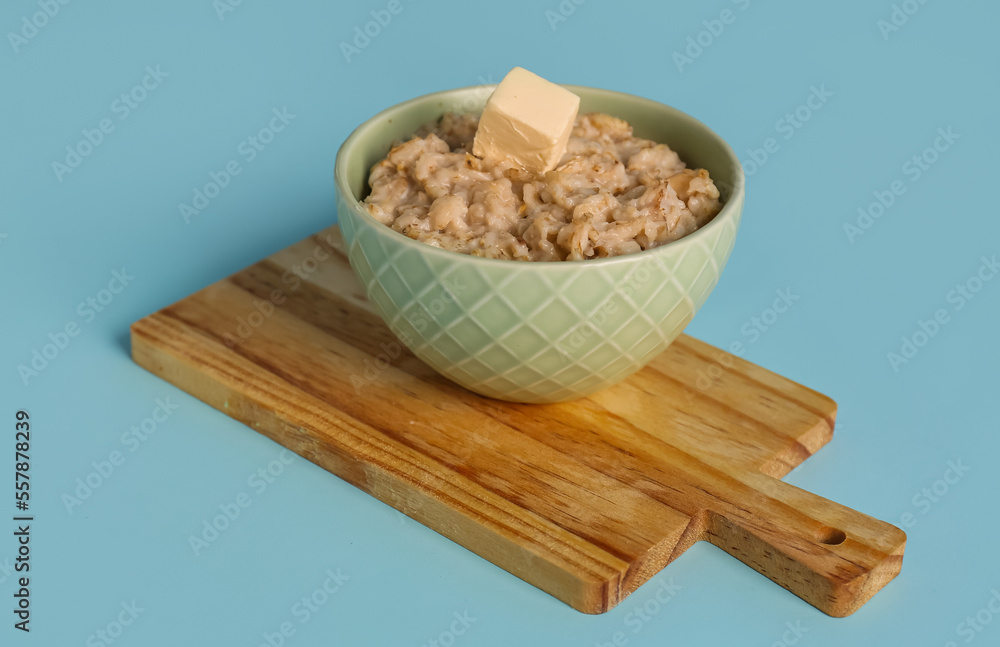 Wooden cutting board, bowl with tasty oatmeal and butter on blue background