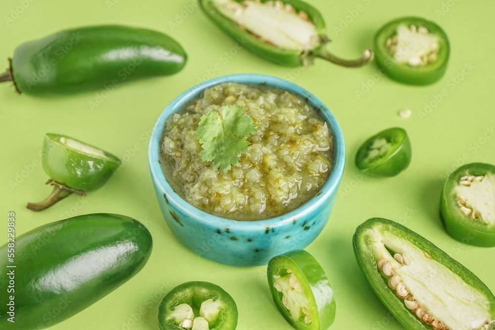 Bowl of tasty green salsa sauce and jalapeno peppers on green background