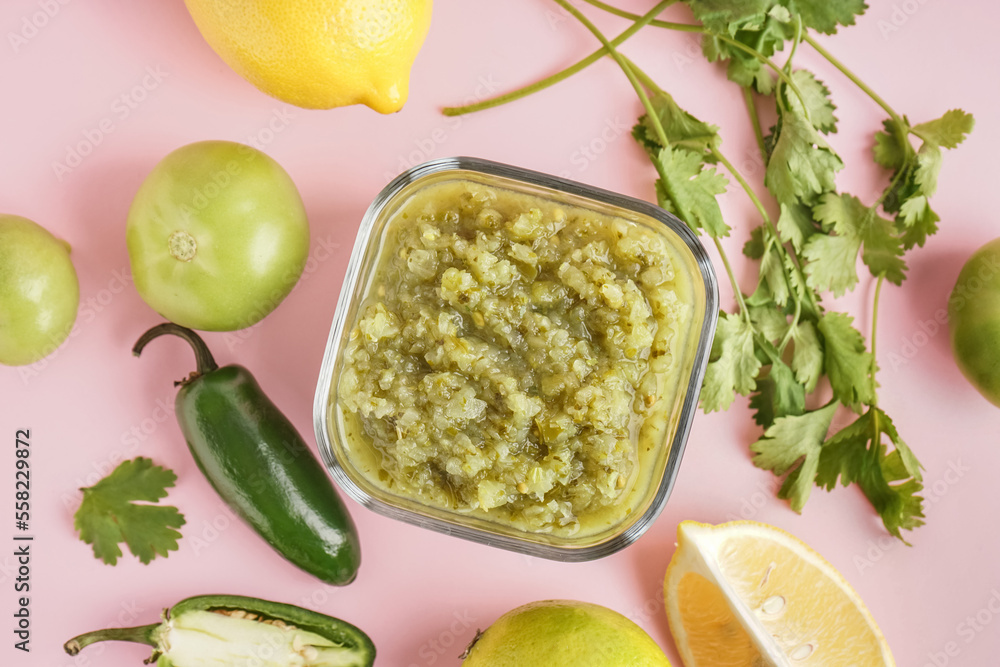 Glass bowl of tasty green salsa sauce and ingredients on pink background