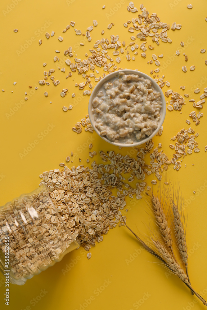 Bowl with tasty oatmeal, overturned jar, heap of flakes and wheat ears on yellow background