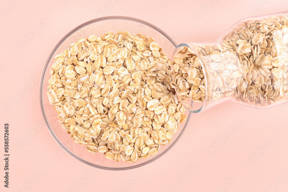 Pouring of raw oatmeal from bottle into bowl on pink background, closeup