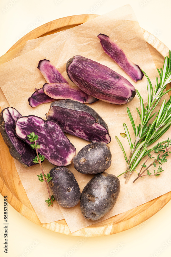 Wooden plate with raw purple potatoes and herbs on color background, closeup