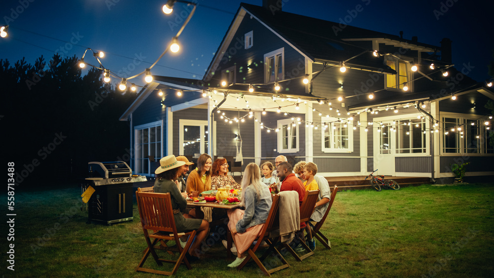 Parents, Children and Friends Gathered at a Barbecue Dinner Table Outside a Beautiful Home with Ligh