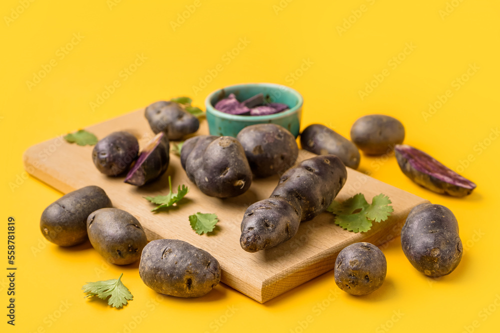 Wooden board with raw purple potatoes and cilantro on color background