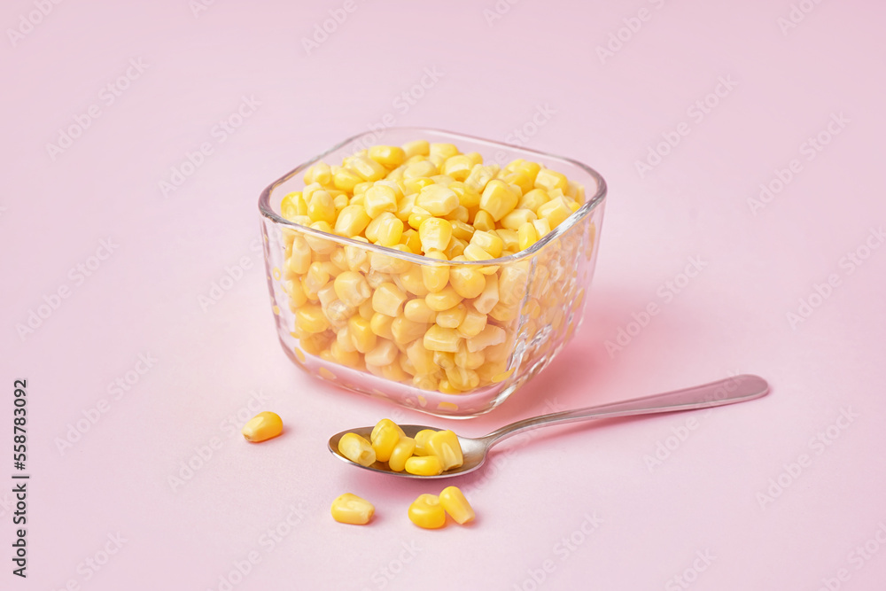 Bowl with canned corn kernels and spoon on pink background