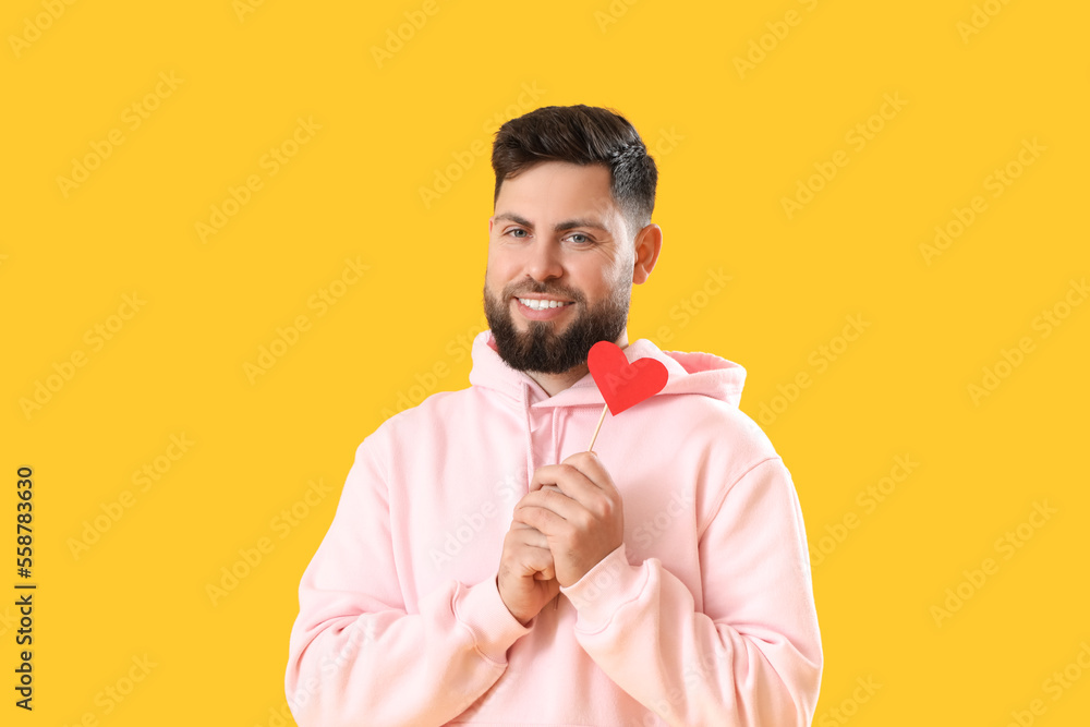 Young bearded man with paper heart for Valentines Day on yellow background