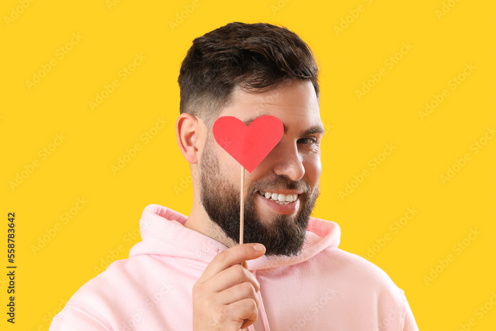 Young bearded man with paper heart for Valentines Day on yellow background, closeup