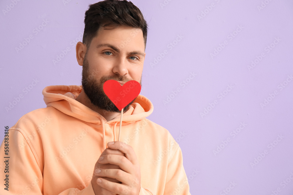 Young bearded man with paper heart for Valentines Day on lilac background, closeup