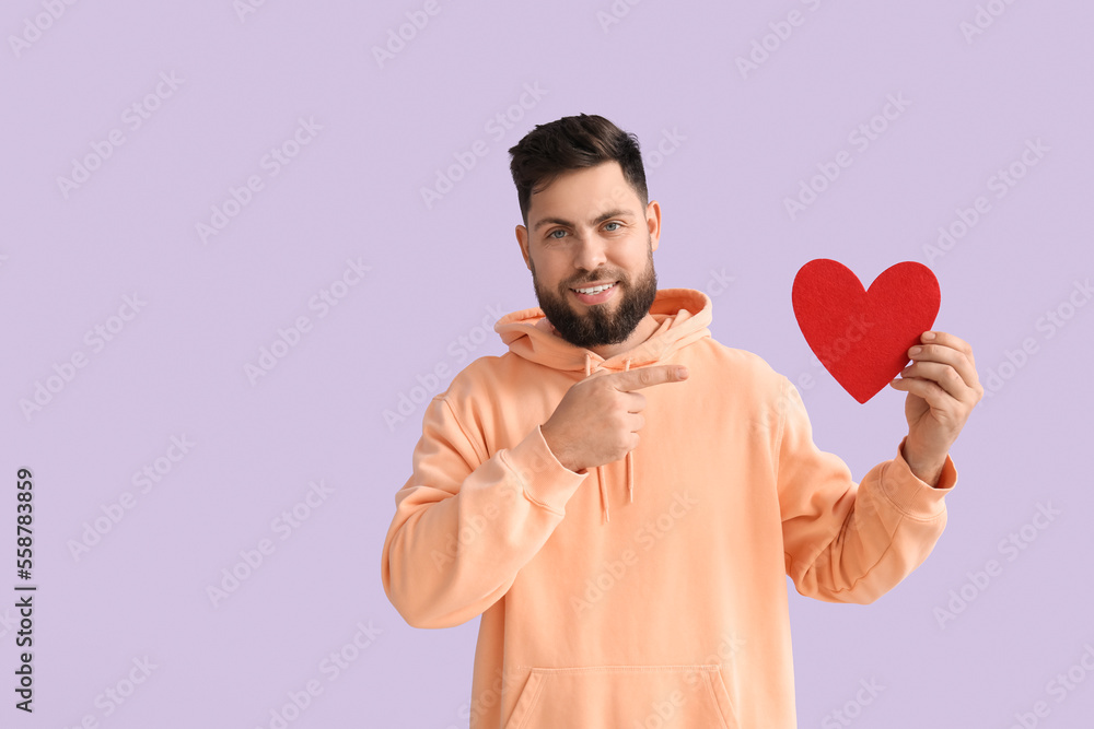 Young bearded man pointing at paper heart for Valentines Day on lilac background