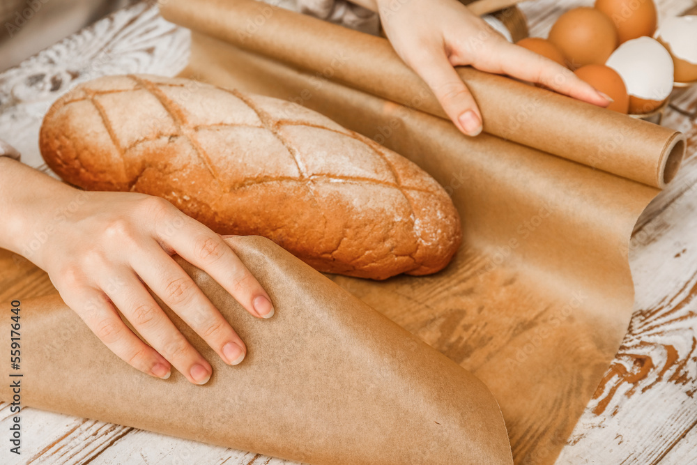 Woman wrapping fresh bread with baking paper on white wooden background, closeup