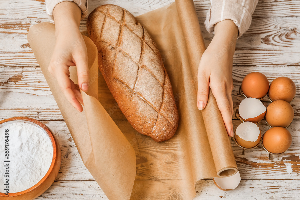 Woman wrapping fresh bread with baking paper on white wooden background, closeup