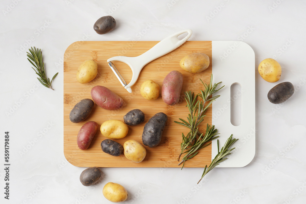 Wooden board with different raw potatoes, rosemary and peeler on light background