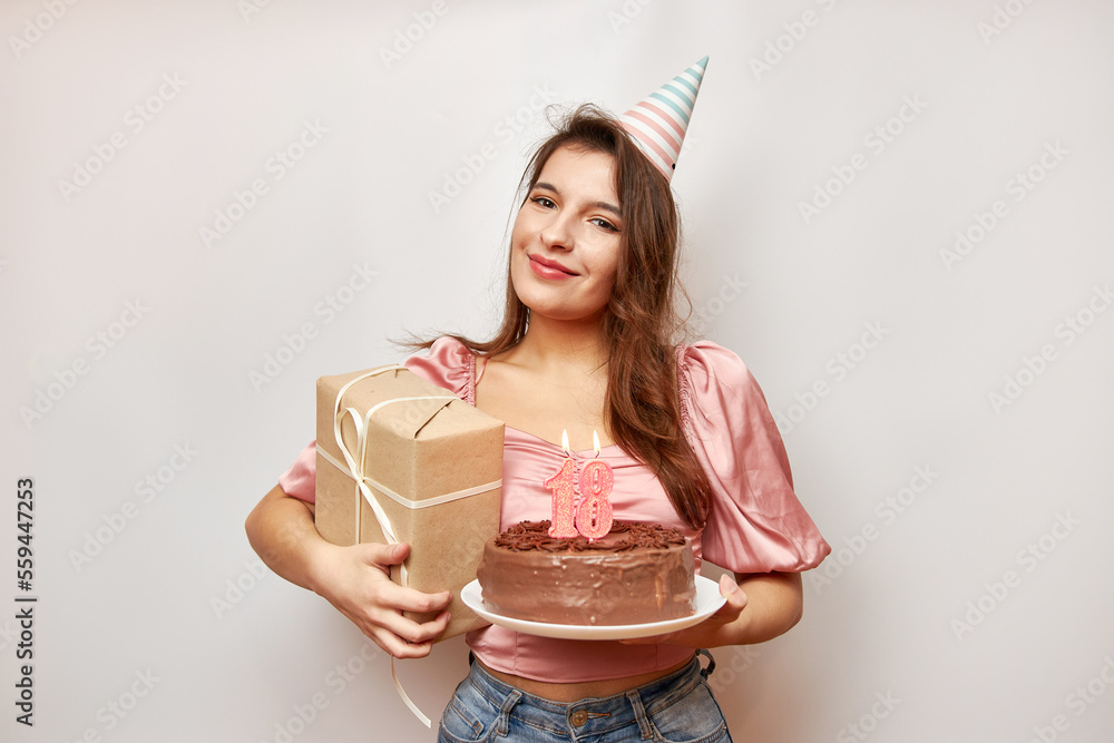 The girl is holding a festive cake with a candle in the form of the number 18 and a gift. Birthday c