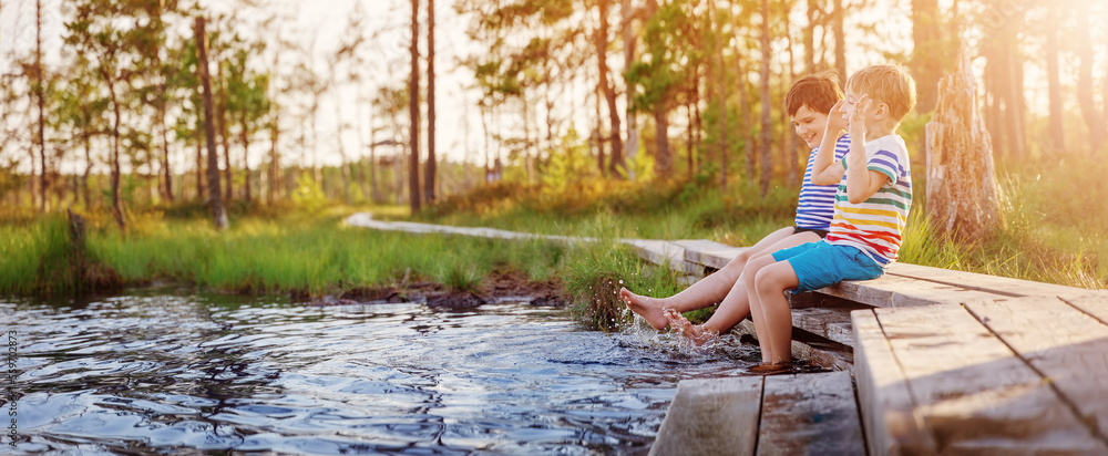 Two boys sitting on the pier of the swamp lake and making splashes by legs.