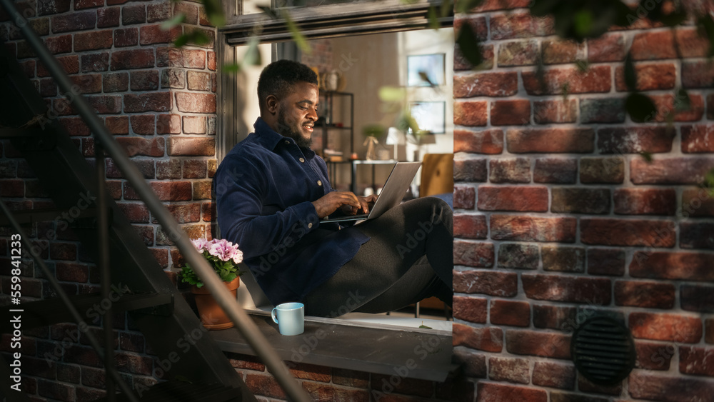 Smiling Black Man Working on Laptop Computer while Sitting on a Windowsill in a Stylish Cozy Brookly