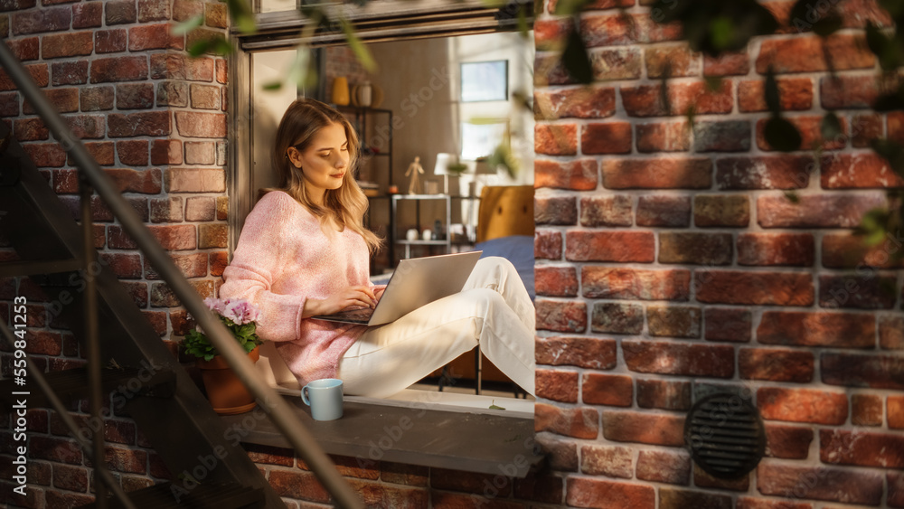 Young Caucasian Woman Working from Home and Using Laptop While Sitting on Bedroom Windowsill on a Wa