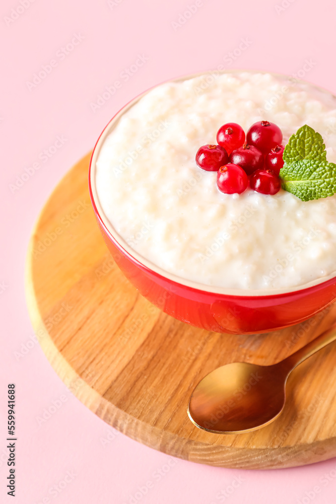 Board with bowl of delicious rice pudding, cranberries, mint leaves and spoon on pink background