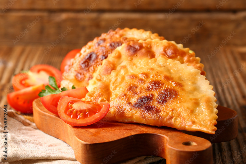 Cutting board with tasty chebureks and tomato slices on wooden background