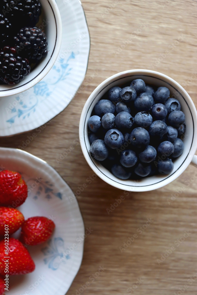 Vintage porcelain cups and plates filled with various fresh berries. Wooden background, top view.