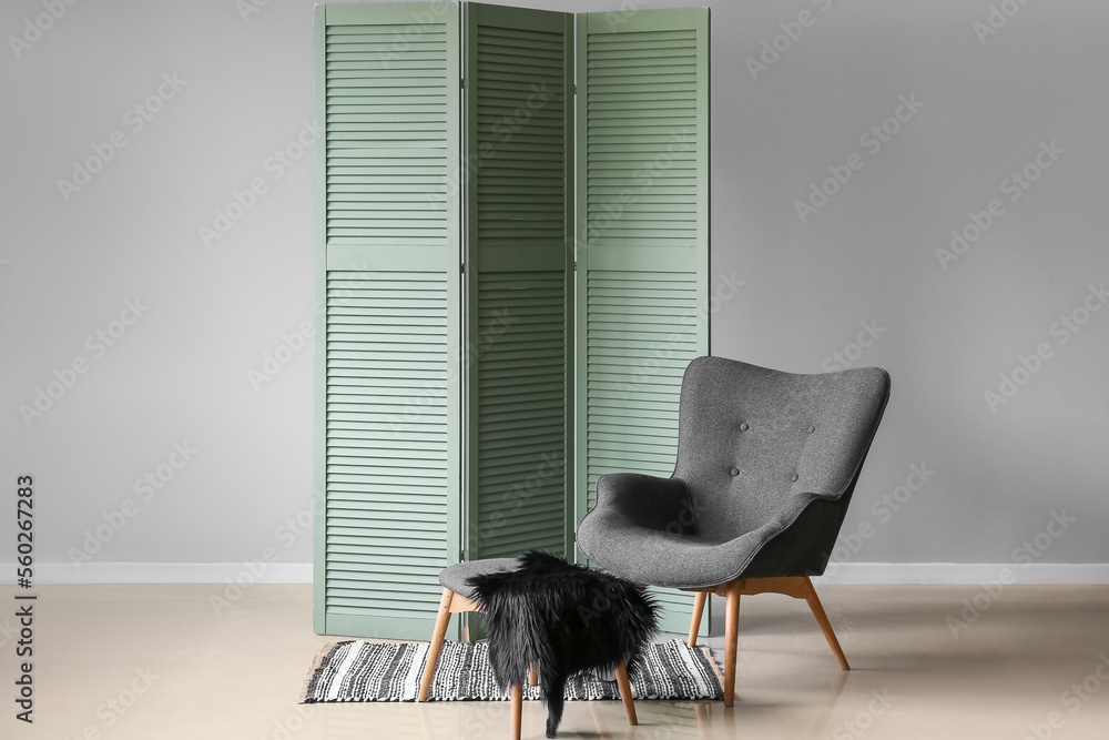 Armchair with ottoman and green folding screen near grey wall